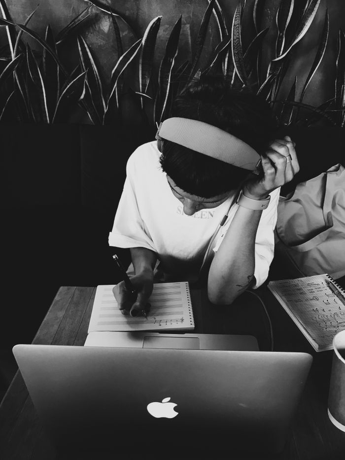 A person writing notes at a MacBook laptop in a monochrome setting, captured in black and white.